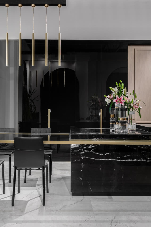 czarne meble kuchenne. Blat szklany ze złotym wykończeniem. Złote lampy wiszące na d stołem. Czarne krzesła.