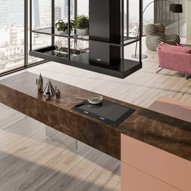 Franke, okap T-Shelf, wyspa kuchenna, płyta grzewcza, patelnia, okap podwieszany z półkami, kwiaty, kieliszki, kanapa, fotel, lampa podłogowa, duże okno, drewniana podłoga