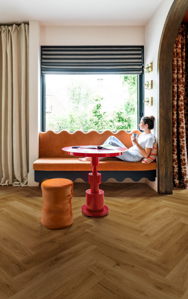 Wnętrze w stylu retro z drewnianą podłogą w jodełkę, pomarańczową ławką przy oknie, czerwonym stolikiem i designerskim stołkiem.