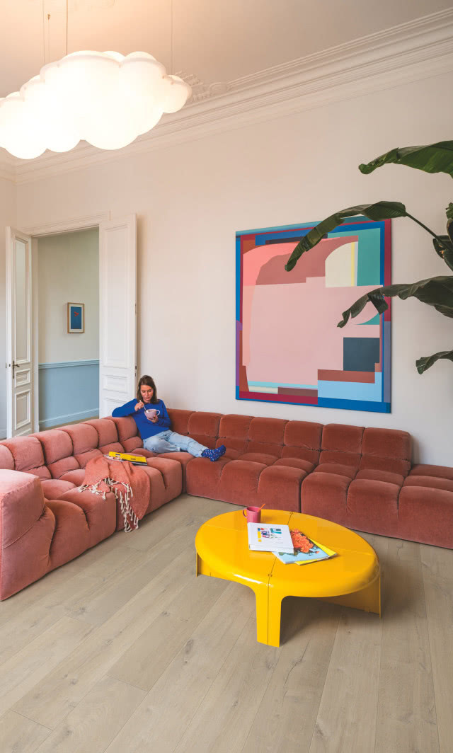 Salon w stylu retro z modułową różową sofą, żółtym stolikiem, kolorowym obrazem i jasną podłogą inspirowaną drewnem.