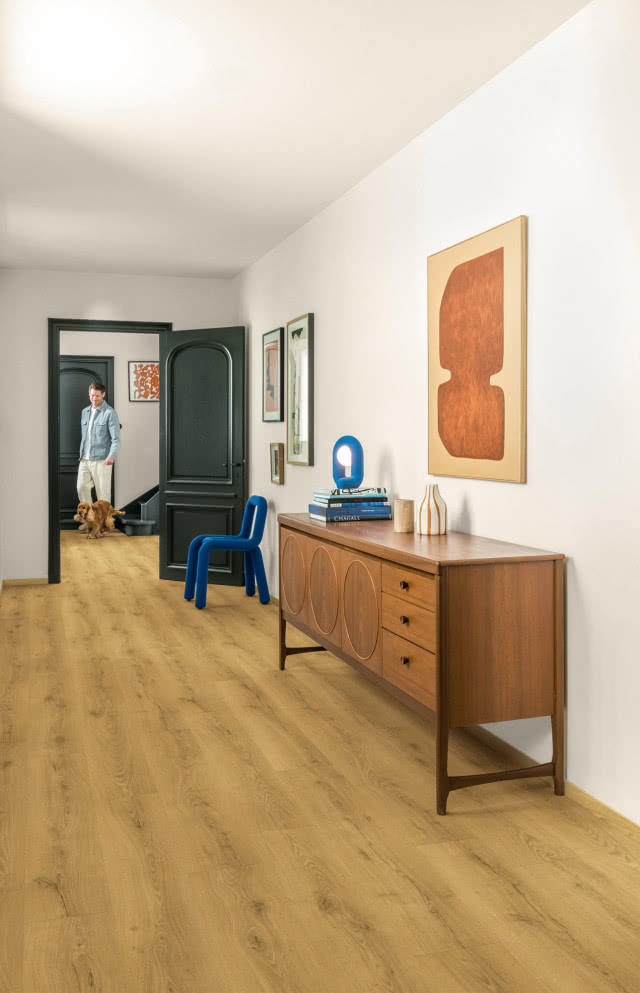 Korytarz w stylu retro z drewnianą komodą vintage, niebieskim krzesłem, obrazami i podłogą inspirowaną naturalnym drewnem.