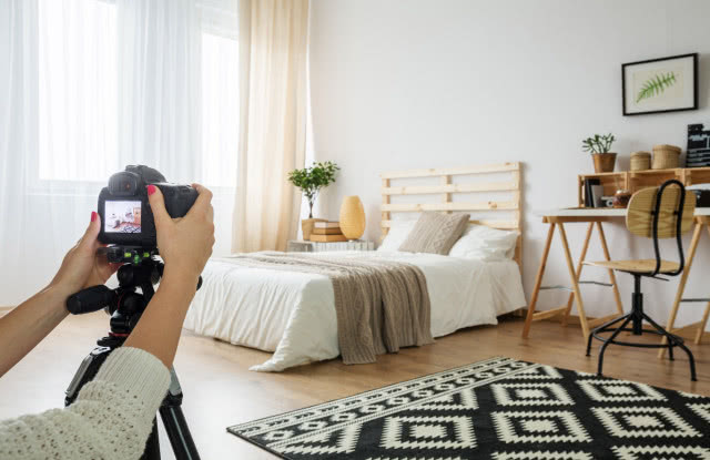 Aparat na statywie fotografuje jasną, minimalistyczną sypialnię z drewnianym łóżkiem, biurkiem i naturalnymi dodatkami.