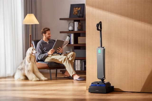 Bezprzewodowy odkurzacz pionowy stojący w stacji dokującej przy ścianie w salonie, w tle mężczyzna czytający magazyn na fotelu obok dużego psa na drewnianej podłodze.
