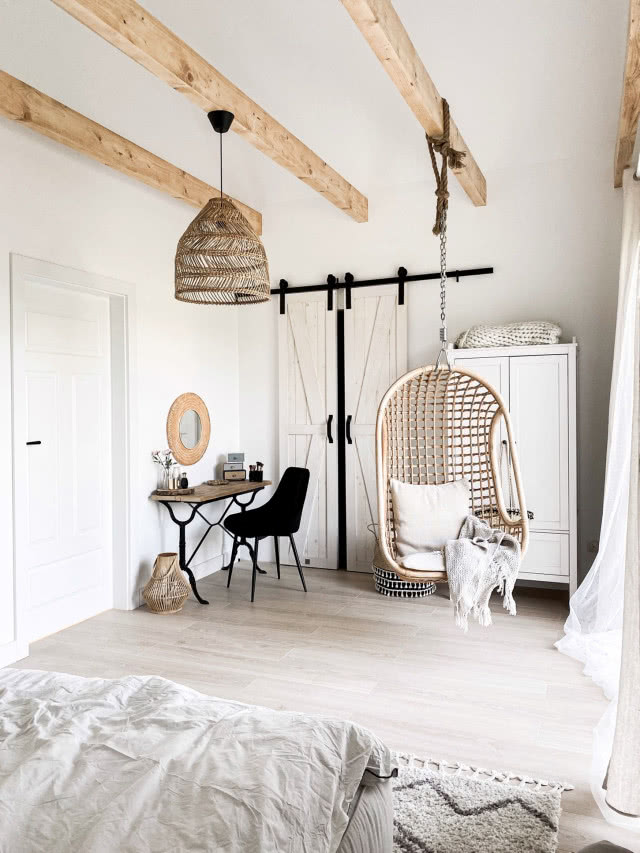 Przytulna sypialnia w stylu boho z rattanową lampą, drewnianymi belkami i wiszącym fotelem w naturalnych kolorach.