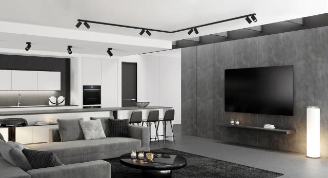szary narożnik, czarny dywan, czarny stolik kawowy, świece, telewizor, ściana imitująca beton, lampa podłogowa, białe meble kuchenne z czarnym blatem, wyspa kuchenna, stołki, piekarnik, lodówka