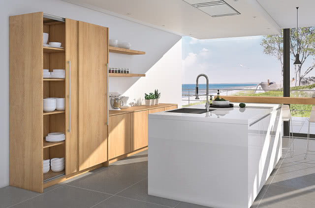 drewniane drzwi przesuwne, wyspa kuchenna w kolorze białym, drewniane fronty kuchenne, drewniany blat kuchenny, półki, czarny zlewozmywak, czarna bateria zlewozmywakowa, płyta indukcyjna, duże okno, jezioro, Morze, drzewa, zastawa stołowa, porcelana