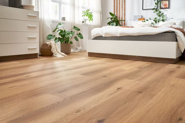 winylowe panele imitujące naturalne drewno, duże łóżko sypialniane, komoda, wiklinowy kosz, duże kwiaty zielone, obrazy, pościel, lamele