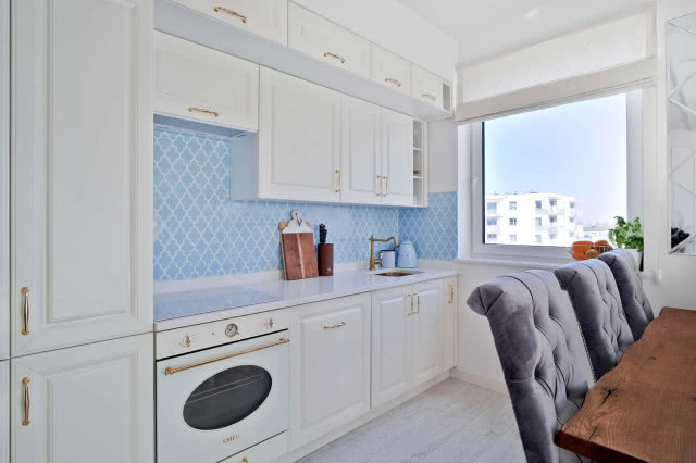 niebieskie płytki, białe meble kuchenne, piekarnik, drewniany stół z krzesłami, duże okno