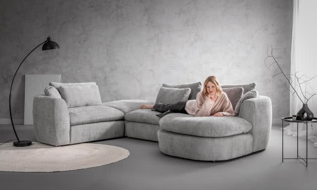 szary narożnik, dywan, lampa podłogowa, stolik kawowy wazon z gałązką, zapachowa świeca, beton na ścianie