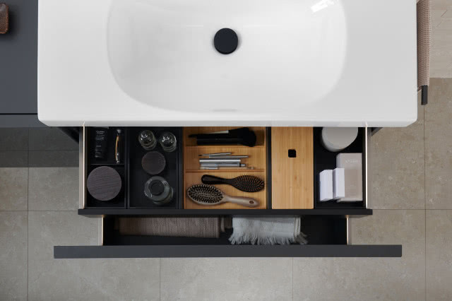 Widok z góry na umywalkę z szafką podumywalkową z otwartą szufladą. We wnętrzu widać przybory kosmetyczne