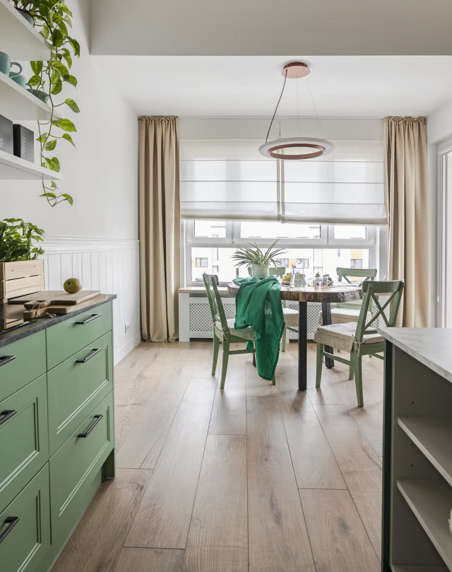 drewniana podłoga, drewniany okrągły stół w jadalni, fotel, krzesła, szafeczka, ekspres do kawy, wyspa kuchenna, zabudowany grzejnik, czarny blat kuchenny, zielone fronty kuchenne