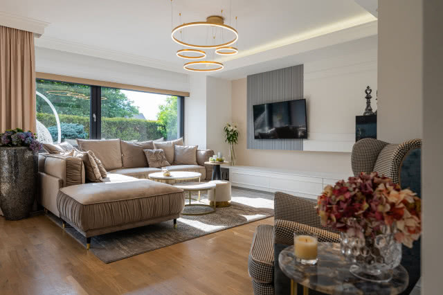 Elegancki salon z jasną sofą narożną, designerskim oświetleniem i dużym oknem wychodzącym na ogród