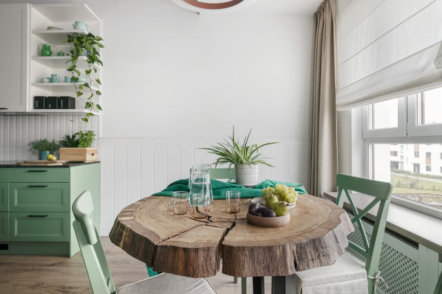 drewniana podłoga, drewniany okrągły stół w jadalni, krzesła, zabudowany grzejnik, czarny blat kuchenny, zielone fronty kuchenne, kwiaty, owoce