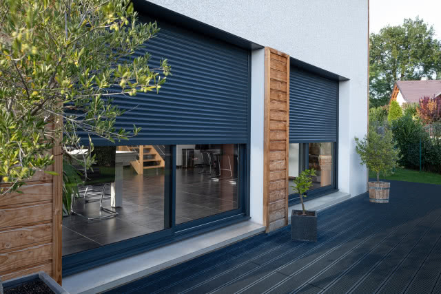 rolety zewnętrzne, drewniana elewacja budynku, taras, donice z drzewkami na tarasie, duże okna, trawa