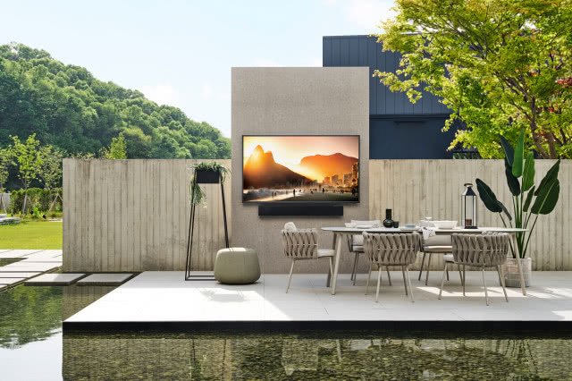 Telewizor wiszący na ścianie w ogrodzie, płot, puf, dwa stoliki okrągłe ogrodowe, krzesła ogrodowe, kwiaty, drzewo, woda