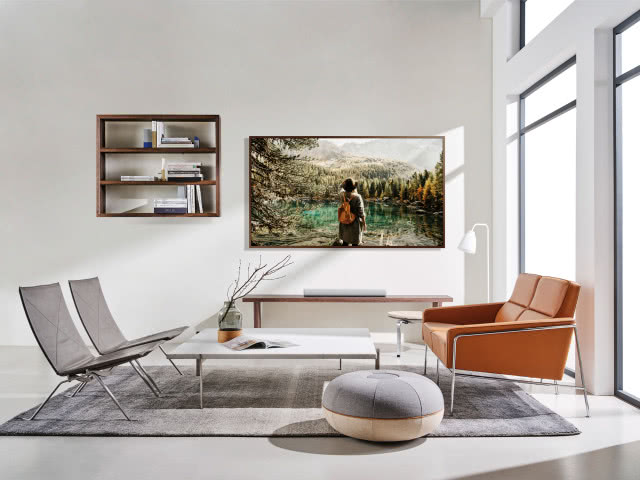 telewizor wiszący na ścianie, projektor na stoliku pod ścianą, dwa szare krzesła, kwadratowy stolik kawowy, pufa, szary dywan, drewniana półka wisząca