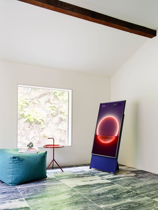 telewizor podobny to smartfona, drewniana belka na suficie, seledynowa pufa, okrągły czerwony stolik