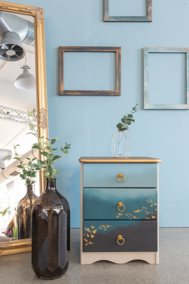 ozdobne ramy na ścianie, duże stojące lustro ze złota rama, cieniowana niebieska komoda z szufladami, złote uchwyty i dekory, duży szklany wazon w kolorze brązowym