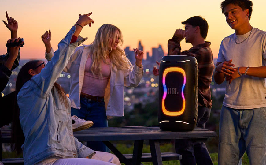JBL PartyBox 130 podczas plenerowej imprezy o zachodzie słońca z efektownym podświetleniem LED i grupą tańczących znajomych.
