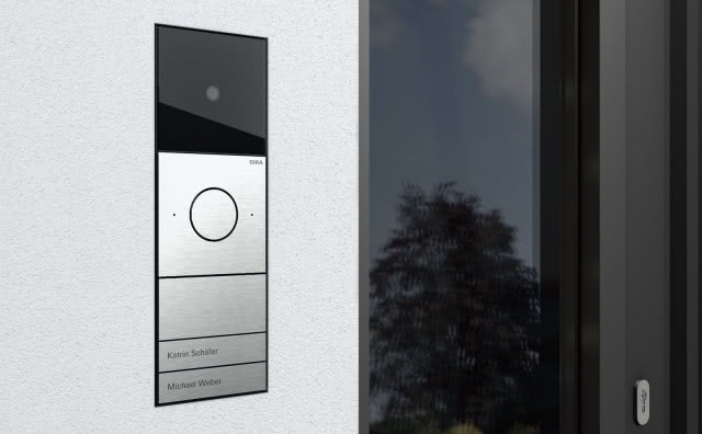Wideodomofon Gira System 106 IP z kamerą i modułowym panelem ze stali szlachetnej, zamontowany na jasnej elewacji obok drzwi wejściowych.