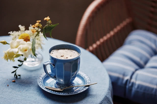 Niebieska porcelanowa filiżanka Rosenthal Maria En Vogue Blue z kawą na spodku, ustawiona na stole obok bukietu kwiatów.