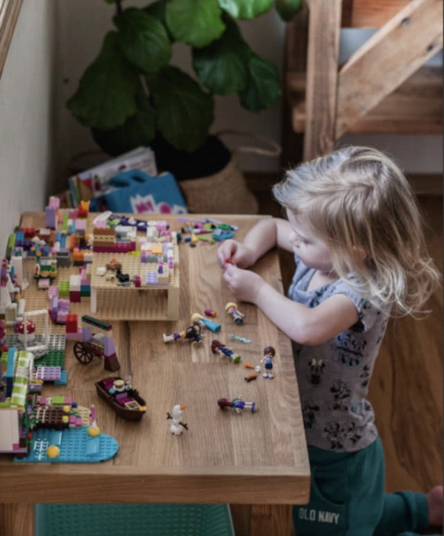Mała dziewczynka bawiąca się klockami LEGO przy drewnianym stole w ciepłym, rodzinnym wnętrzu.