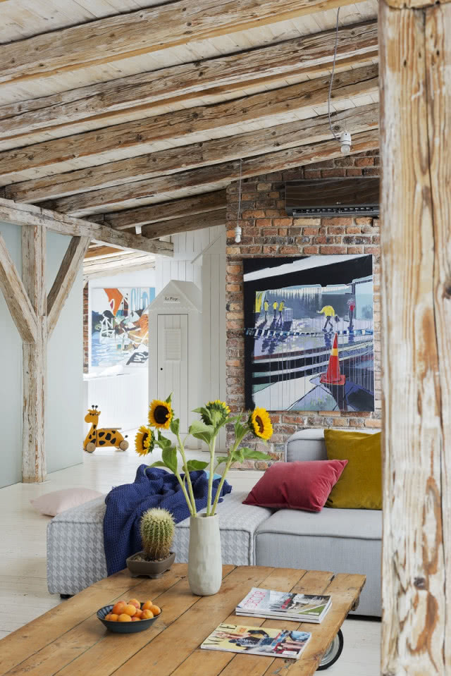 Świeże kwiaty i kolorowe detale we wnętrzu, drewniane belki, słoneczniki, granatowy pled i szara sofa