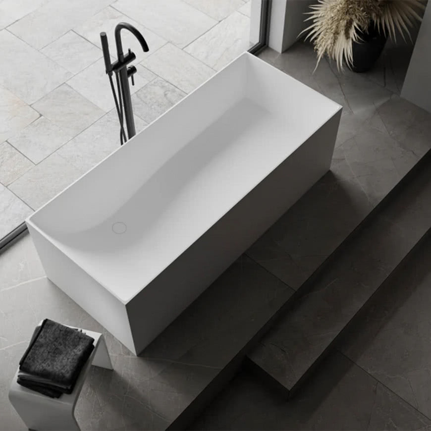 Minimalistyczna, wolnostojąca wanna o prostokątnej formie w jasnym wykończeniu, ustawiona przy oknie, z czarną baterią podłogową na tle kamiennej posadzki.