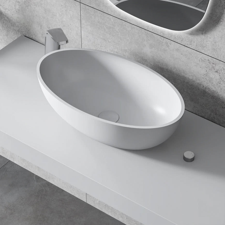 Biała, owalna umywalka nablatowa o miękkich liniach na minimalistycznym blacie, na tle szarej ściany i podświetlanego lustra.