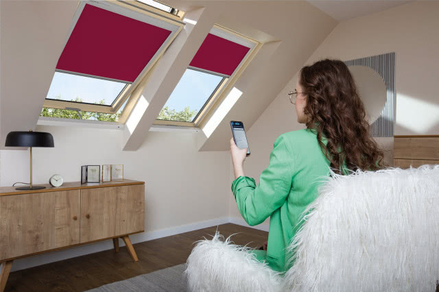 Rolety ARF dostępne są w różnych kolorach, w tym Viva Magenta - kolorze roku 2023 (fot. FAKRO), Kobieta siedzi na fotelu i steruje aplikacją w telefonie roletami, komoda, lampka biurkowa, budzik, okna dachowe