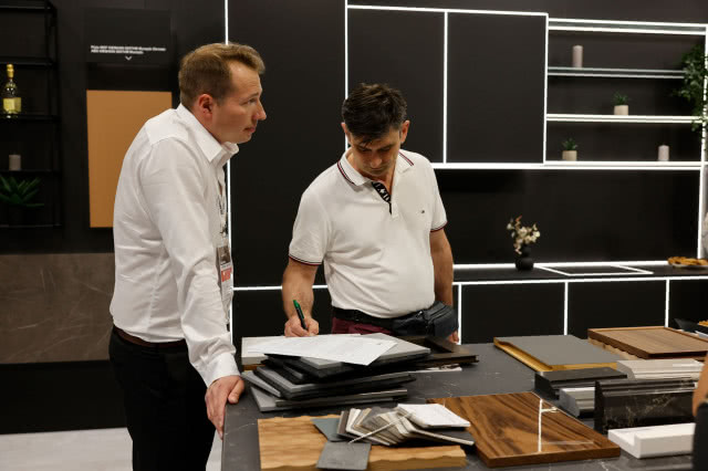 Przedstawiciel firmy omawia ofertę i podpisywanie dokumentów z klientem przy stoisku z próbkami materiałów meblowych podczas targów