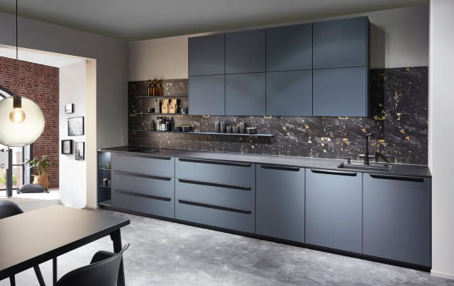 granatowe meble kuchenne, czerwona cegła na ścianie, półki, wiszące szafki, czarne uchwyty, czarna bateria zlewozmywakowa, stół z krzesłami, obrazy, oświetlenie sufitowe