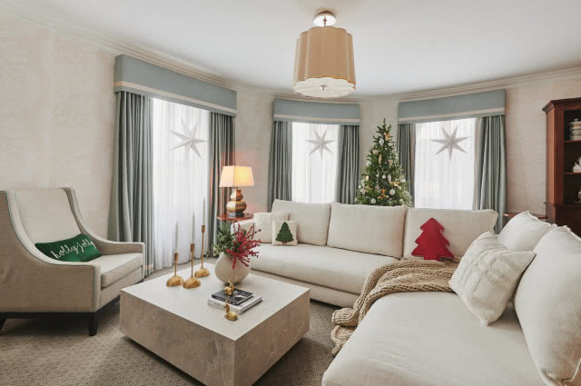 biały narożnik, stolik kawowy, oświetlenie sufitowe, lampy biurkowe, zasony, firany, gwiazdy na oknie, drewniana witryna na bibeloty, obrazy, choinki, poduszki ozdobne, koc, fotel