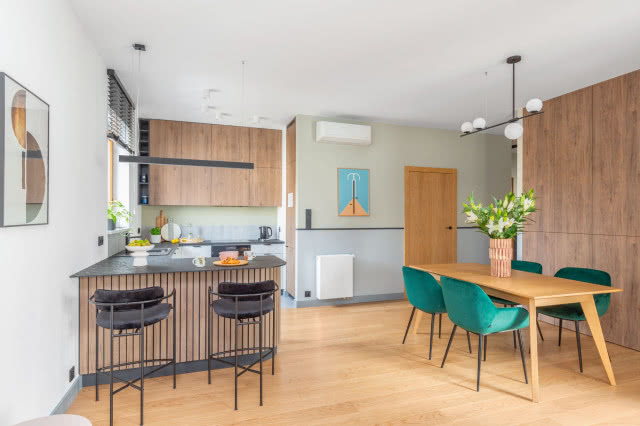 Aleksandra Kurowska postawiła na sprawdzone motywy dekoracyjne. Ta sama zieleń, którą zastosowano w korytarzu,  pojawia się nad blatem roboczym w kuchni, nad narożnikiem w części wypoczynkowej, a także w sypialni. Uwagę zwraca  dekoracyjny pasek w kolorze antracytowym, takim samym w jakim są żaluzje.