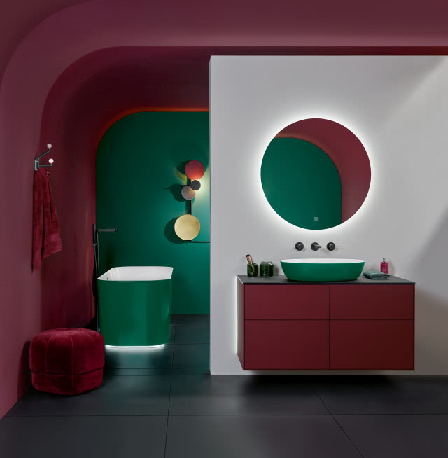 wanna wolnostojąca w kolorze zielonym, umywalka nablatowa zielona, meble łazienkowe w kolorze bordo, pufa, ściany zielono bordowe, okrągłe lustro, ręcznik