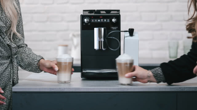 czarny ekspres do kawy, szary blat kuchenny, biała cegłą na ścianie, w szklankach dwie kawy ze spienionym mlekiem