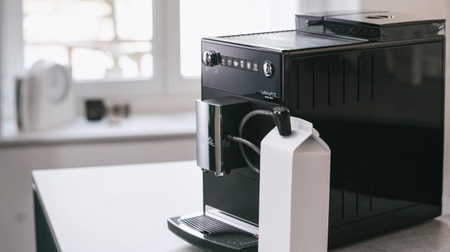 czarny ekspres do kawy, szary blat kuchenny, biała cegłą na ścianie, biały budzik, okno