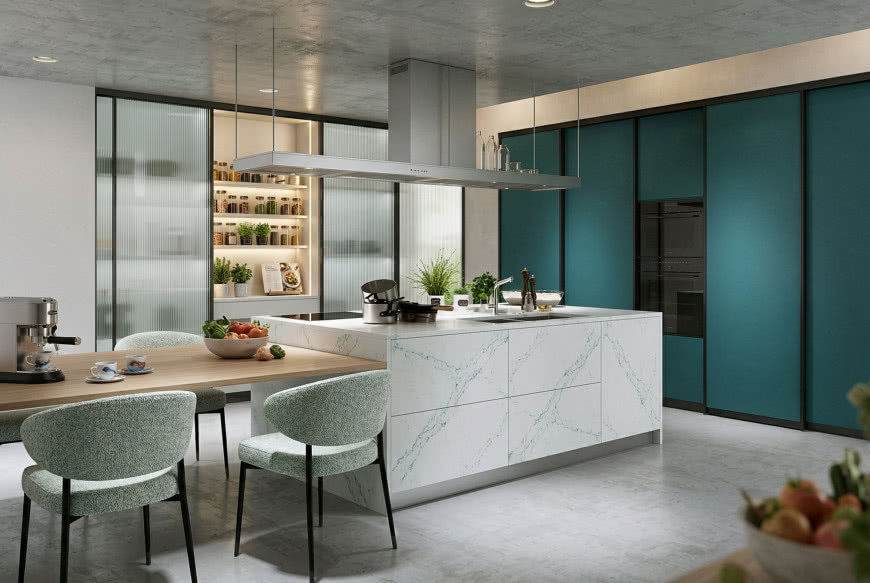Nowoczesna kuchnia z wyspą wykończoną jasnym spiekiem, zielonymi frontami przesuwnymi i przeszkloną spiżarką z podświetlanymi półkami, połączona z drewnianym stołem jadalnianym.