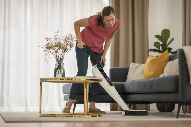 Kobieta odkurza dywan, stolik kawowy, kanapa, poduszki ozdobne, białe firany, beżowe zasłony, wazon z kwiatami, 