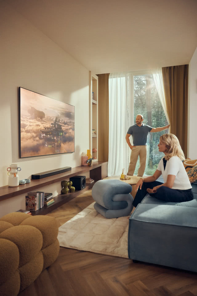 Nowoczesny salon z telewizorem Samsung, soundbarem i dużymi oknami, w którym domownicy odpoczywają przed ekranem.
